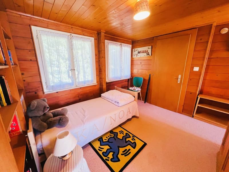 small room with bed, teddy bear, small carpet, chair, small nightstand, shelf, window with white curtains, wooden wall, and lamp