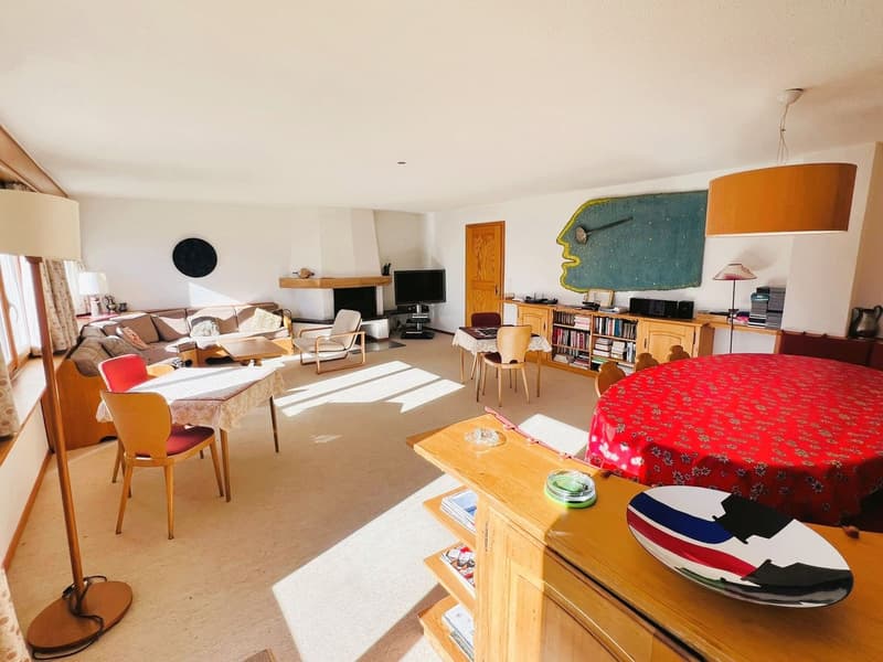 A room with a fireplace, a sofa, a dining table, chairs, a desk, and books on a shelf