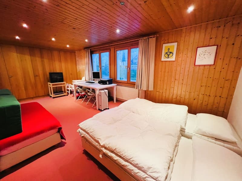 A room with a wooden wall and a desk with a computer monitor. A red carpeted floor and three beds with white covers. There is also a black television on a shelf.