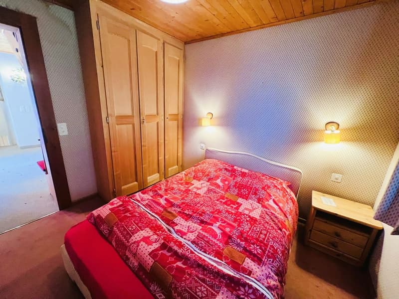 A cozy bedroom with wooden floors, a wooden wardrobe, a bed with a red quilt, a nightstand, and two wall lamps.