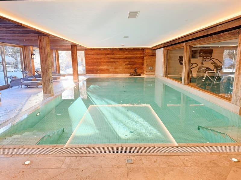 An indoor swimming pool with green water, surrounded by marble tiles and wooden walls. The pool has a shallow section on one side and a deep section in the middle. There are lounge chairs and a table with umbrellas in the background, along with exercise e