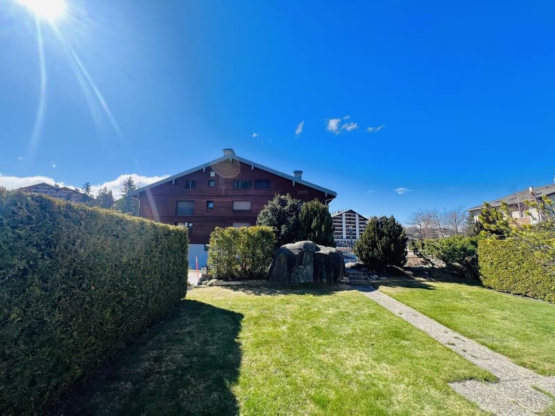 Wooden house, multiple windows, well-maintained garden, hedges, pathway, large rock, clear blue sky