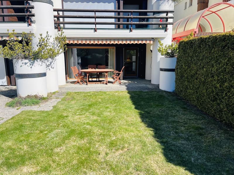 A small garden with a table and chairs, a balcony, and a hedge.