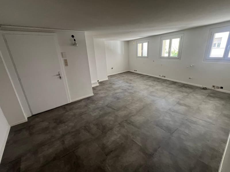 Large open-plan living room with white walls, concrete-look flooring, and two windows providing natural light.