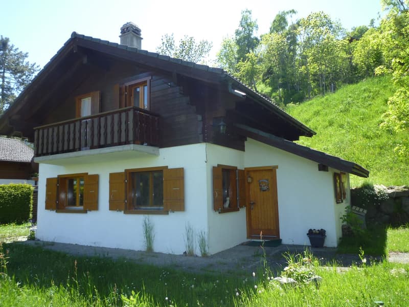 Two-story wooden chalet-style house with a balcony, surrounded by lush greenery and trees