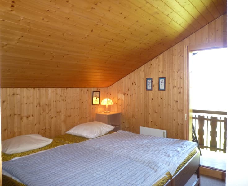 Cozy wooden bedroom with a double bed, bedside table with a lamp, and a balcony or terrace visible through the window.