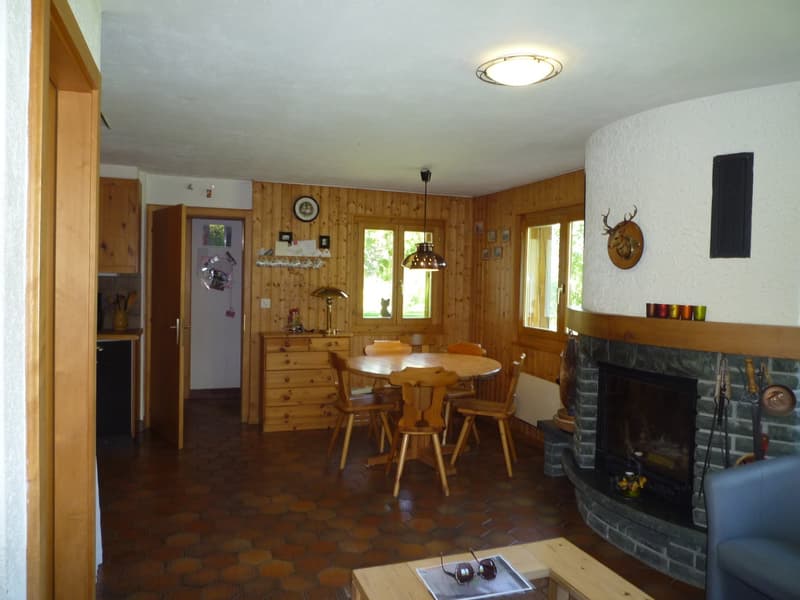The image shows a cozy, rustic-style living and dining area. The room has wood-paneled walls, a round wooden dining table with chairs, and a fireplace with a stone surround. There are various decorative elements such as a mounted deer head, framed artwork