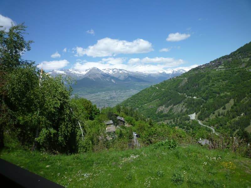The image shows a scenic mountain landscape with snow-capped peaks in the distance, surrounded by lush green forests and meadows. There are small buildings or cabins visible in the valley below, and a winding road can be seen snaking through the landscape