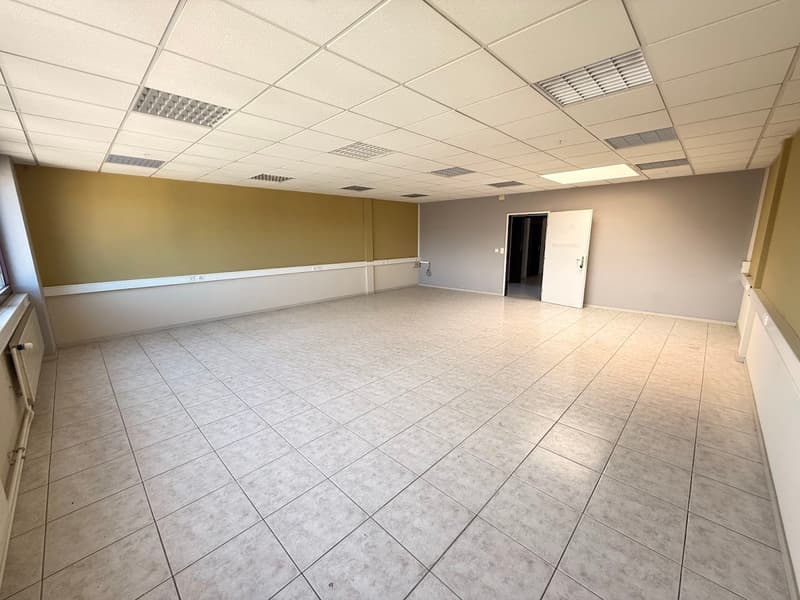 The image shows a large, open office space with a tiled floor and a suspended ceiling with recessed lighting. The walls are painted in a neutral color, and the room appears to be empty, ready for use as an office or commercial space.