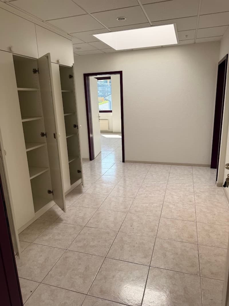 The image shows a spacious office space with a tiled floor, white walls, and recessed lighting. There are built-in shelves and cabinets along one wall, and a doorway leading to another room is visible in the background.