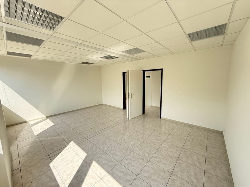 The image shows an empty office space with a tiled floor, white walls, and a suspended ceiling with recessed lighting. The room has two doors, one of which is visible. The space appears to be a commercial or office environment.