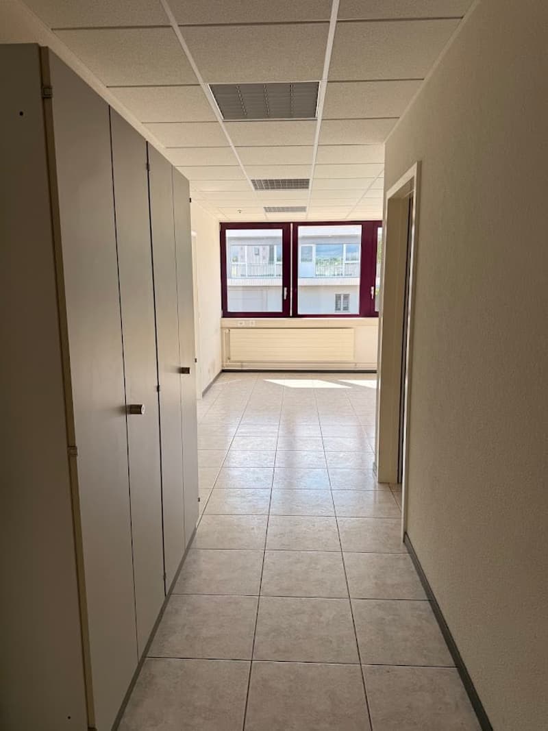 The image shows a hallway or corridor in an office or commercial building. The space has a tiled floor, white walls, and a suspended ceiling with recessed lighting. There are several closed doors along the hallway, suggesting individual office or work spa