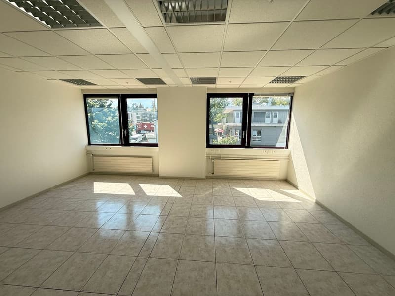The image shows an empty office space with a tiled floor, suspended ceiling, and large windows overlooking the exterior. The room appears to be a commercial or office space, with ample natural light coming in through the windows.