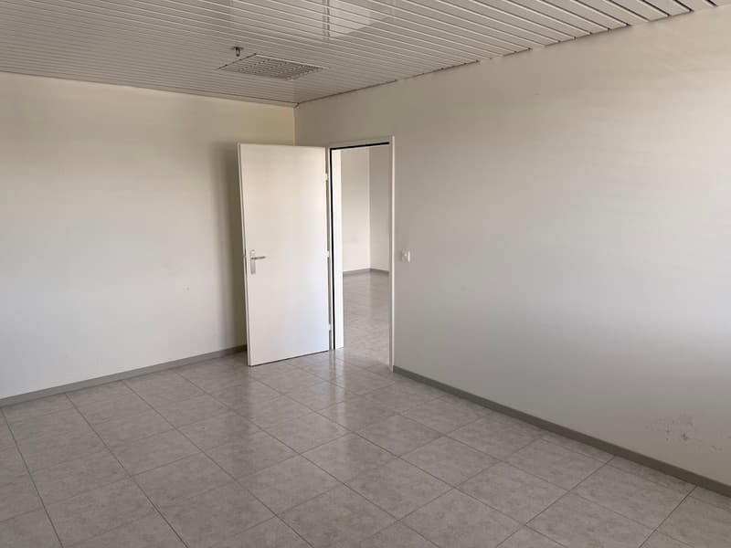The image shows an empty room with white walls, a tiled floor, and a suspended ceiling. There are two white doors visible, one of which is open, leading to another room. The room appears to be a simple, unfinished space, likely intended for commercial or