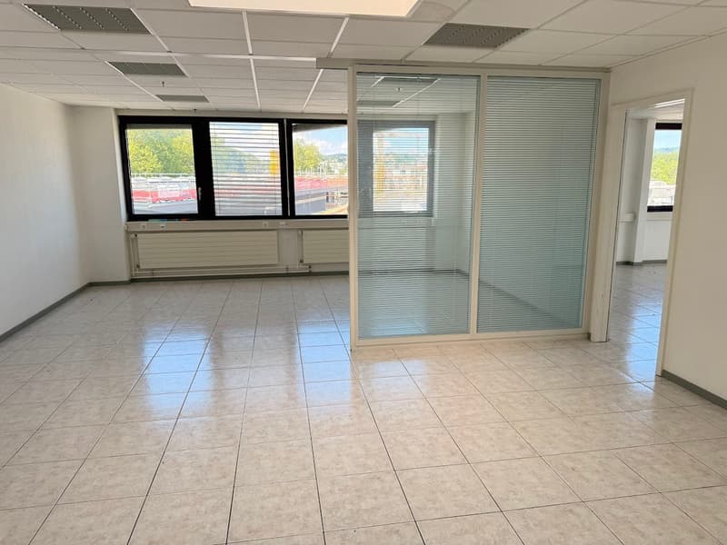 The image shows a spacious office space with large windows, a tiled floor, and a glass partition wall. The room appears to be well-lit and has a modern, professional atmosphere.