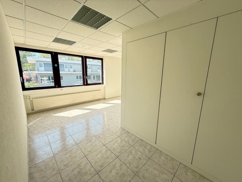 The image shows an empty office space with a tiled floor, white walls, and large windows overlooking the exterior of the building. The room has a suspended ceiling with recessed lighting fixtures. There is a closed door visible on the right side of the ro