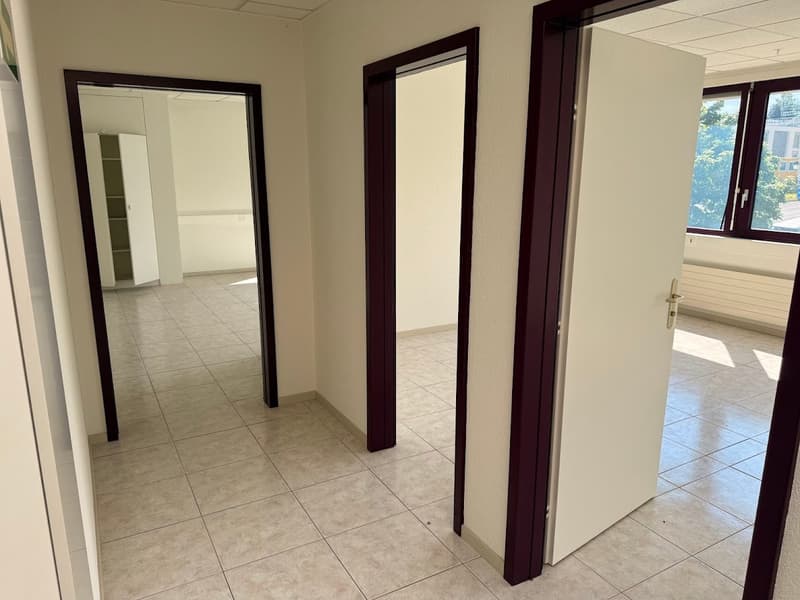 The image shows an empty office space with tile flooring, white walls, and large windows. There are two dark-framed mirrors on the walls, and a door leading to another room. The space appears to be a commercial or office setting.