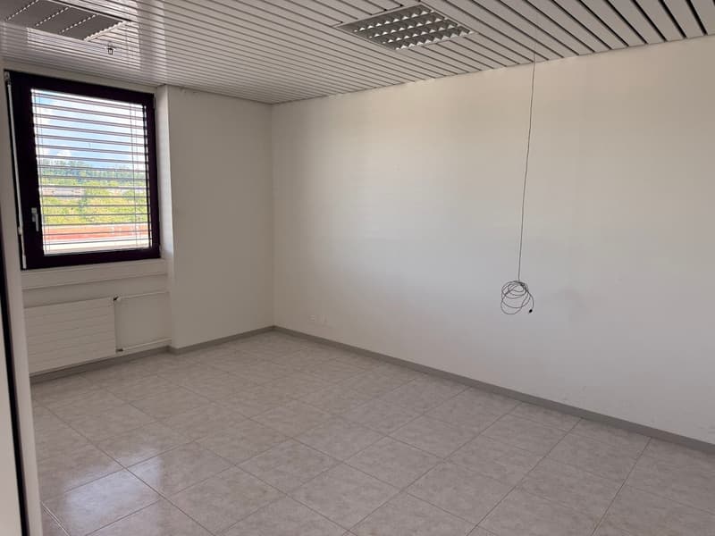 The image shows an empty office space with white walls, a tiled floor, and a window overlooking the outdoors. The ceiling has a grid-like pattern and there is a light fixture hanging from the ceiling.