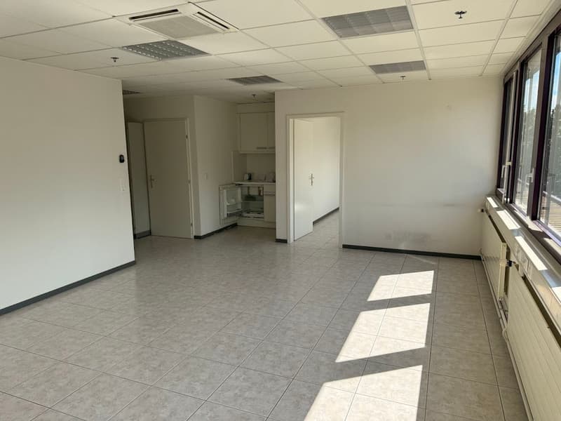 The image shows a large, open office space with a tiled floor, white walls, and a kitchenette area visible in the background. The room has a clean, minimalist appearance with ceiling-mounted air conditioning units and large windows providing natural light
