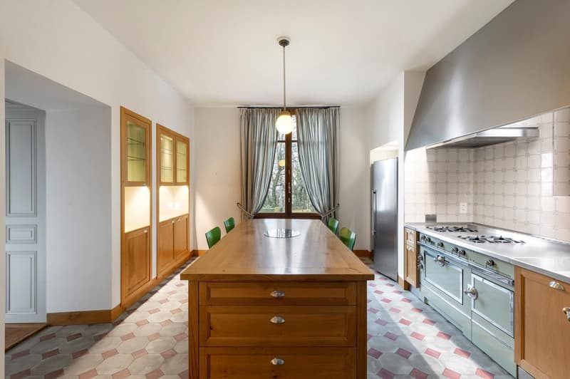 large wooden table, green chairs, large window with curtains, pendant light, stainless steel refrigerator, stove with chimney, wooden cabinets