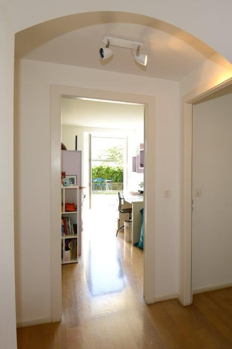 Wooden floor, white walls, open entrance to another room with desk and chair, cabinet with books and items