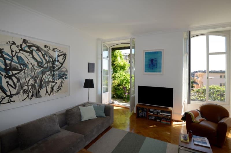 living room with wooden floor, grey sofa, large window leading to balcony, TV, artwork on the wall