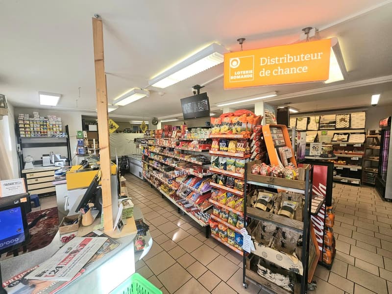 A store interior with various shelves stocked with snacks, drinks, and other items. A counter with magazines and newspapers. A sign reading 'Distributeur de chance' hangs from the ceiling.