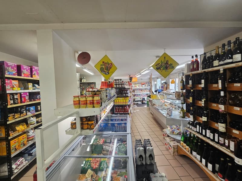 Interior of a store with various products on shelves, including snacks, beverages, and condiments. A refrigerated section with food items and a display case with bottles. Signs and ceiling lights are visible.