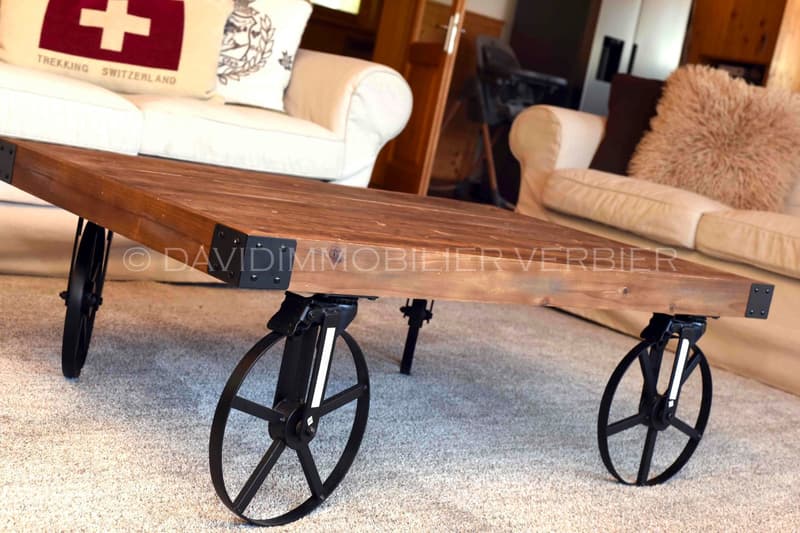 wooden coffee table with metal wheels, positioned between two sofas, a large white throw pillow, a red pillow with Swiss cross on it