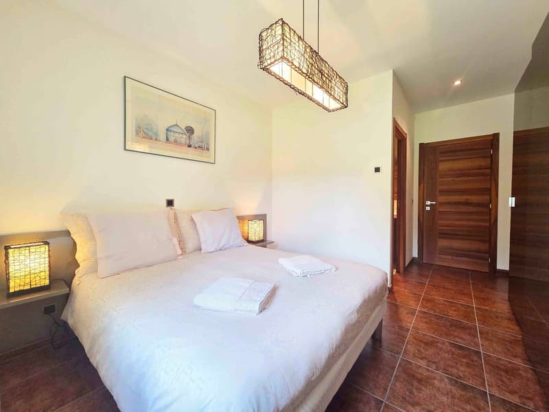 A spacious bedroom with white bedding, wooden headboard, a chandelier, wooden bedside tables, lamps, a framed picture on the wall, and brown tiled flooring.
