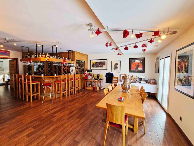 a room with a bar, wooden flooring, wooden dining table and chairs, decorative wine glasses on the bar, a silver pitcher on the table, wall decorations