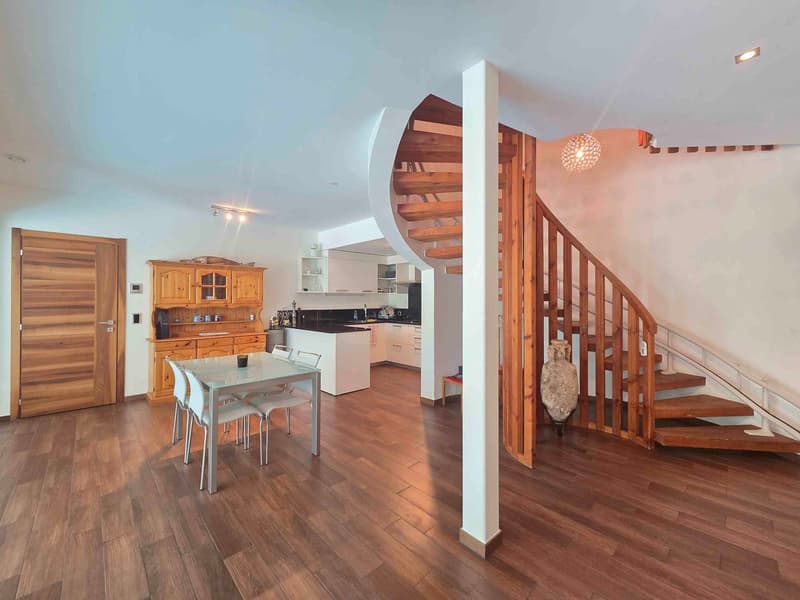 Dining area with wooden floor, spiral staircase, dining table, chairs, kitchen with modern cabinets, black countertop, kitchen appliances, wooden cabinet with glass doors, ceiling lamps