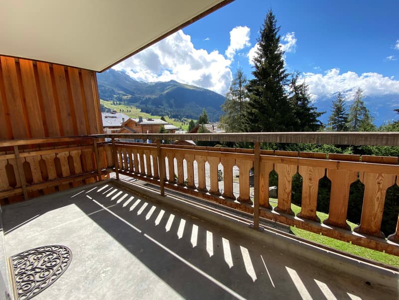 Balcony with wooden railings, wooden slats, concrete floor, outdoor area with grass, mountain range, sky with clouds, greenery