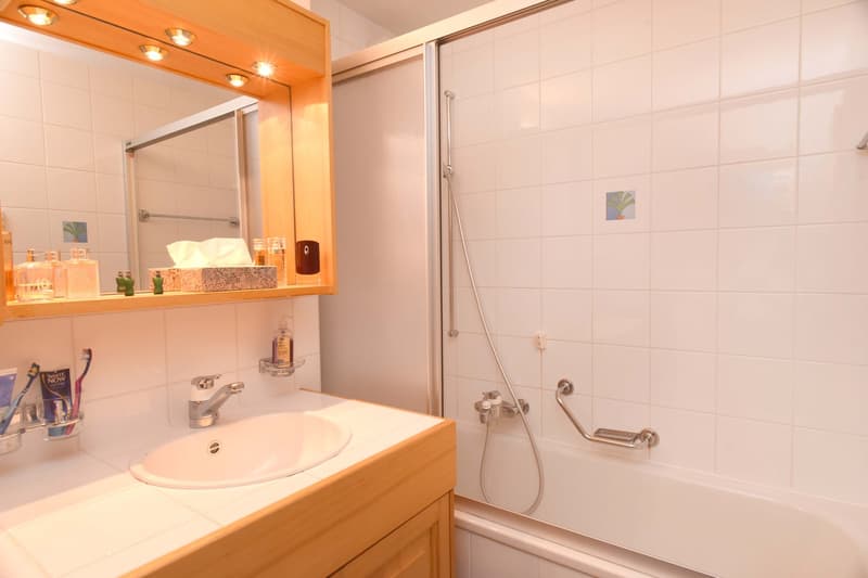 white tiled walls, silver metal faucet, sink with toothbrushes, walk-in shower, white bathtub, mirror