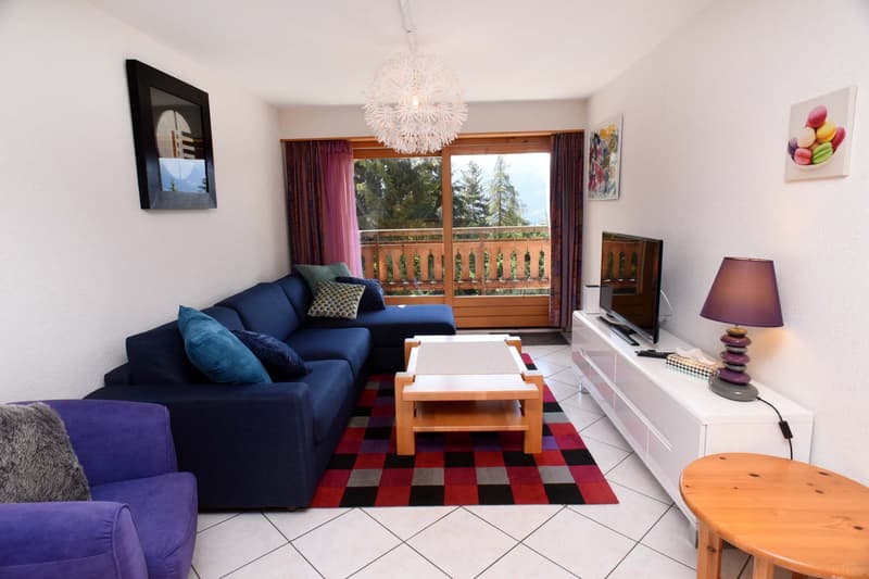 Blue sectional sofa, wooden coffee table, flat screen TV, white tile flooring, decorative rug, wooden side table, ceiling lamp, wooden balcony railings