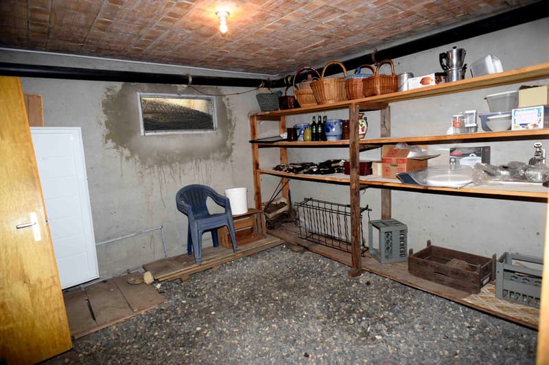 A storage room with wooden shelves, baskets, and various items on the shelves. A small chair, a white bucket, and a white door are present. There is a brick ceiling, and an open window is on the wall.
