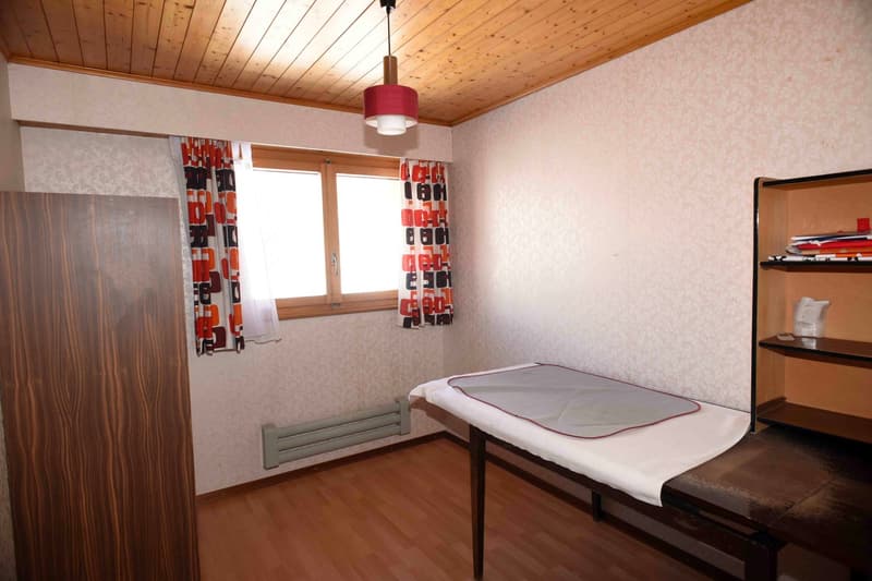 Single bed, wooden floor, white walls, window with curtains, small lamp, wooden cabinet, shelf