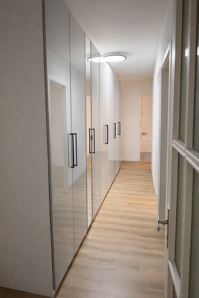 White walls, wood floor, large mirrored wardrobe, lighted hallway, bathroom