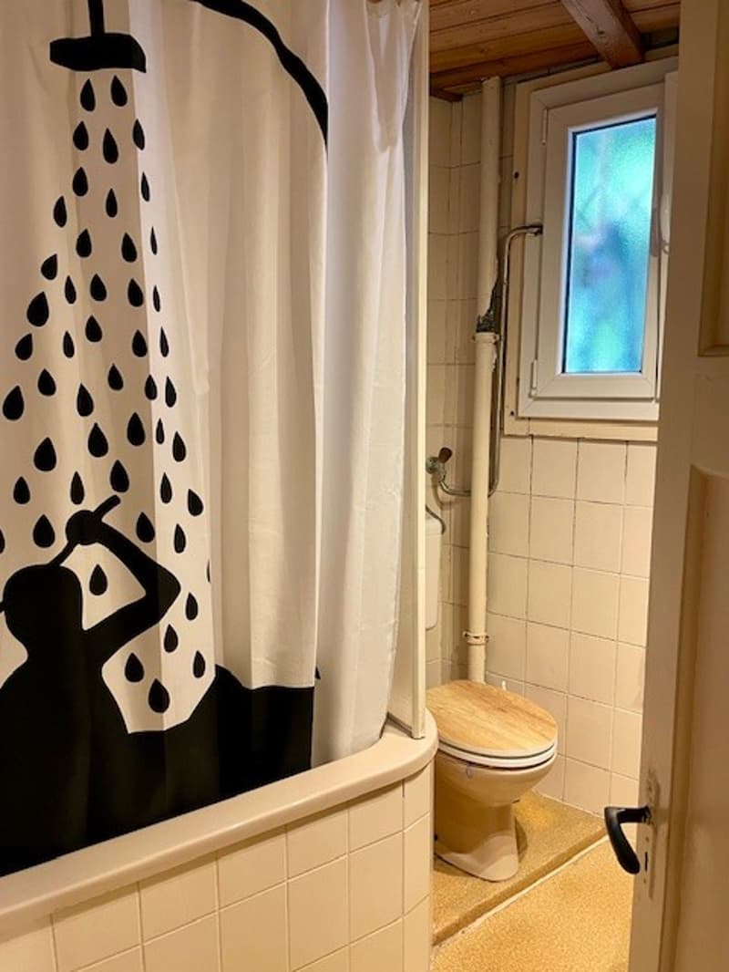 Bathroom with bathtub and toilet, shower curtain with black silhouette and water droplet design, beige tiled walls and floor, wooden ceiling, small window, white door handle
