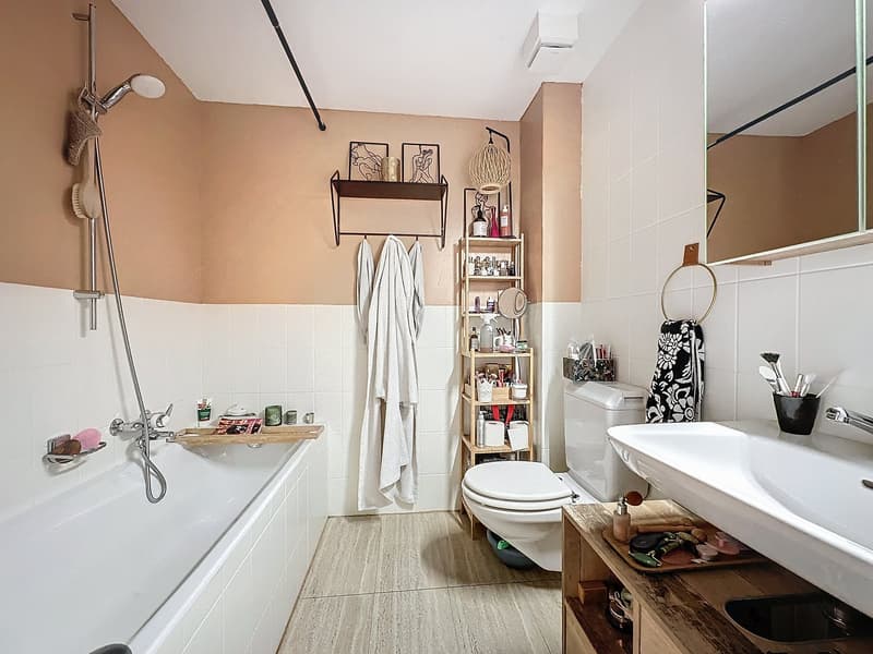 Modern bathroom with white tiles, a bathtub, shower, white sink, toilet, towels, showerhead, and small wooden cabinet with storage shelves.