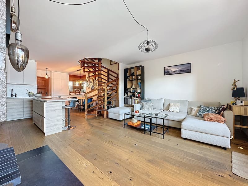Wooden flooring, open kitchen with dining area, central staircase, living room with large sofa and armchairs, desk with picture, fireplace, wooden shelves