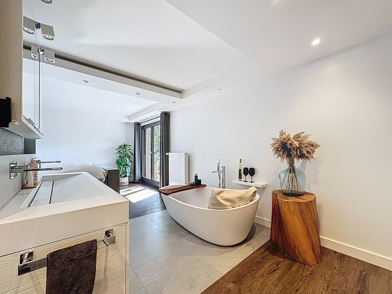 White bathtub with tray, wooden side table, large white sink, towel, potted plant, double glass door with window.