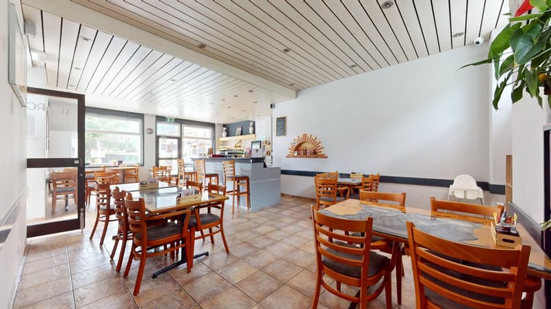 Interior of restaurant with wooden tables, tiled floor, white walls, windows, counter, shelves with bottles, menu board.