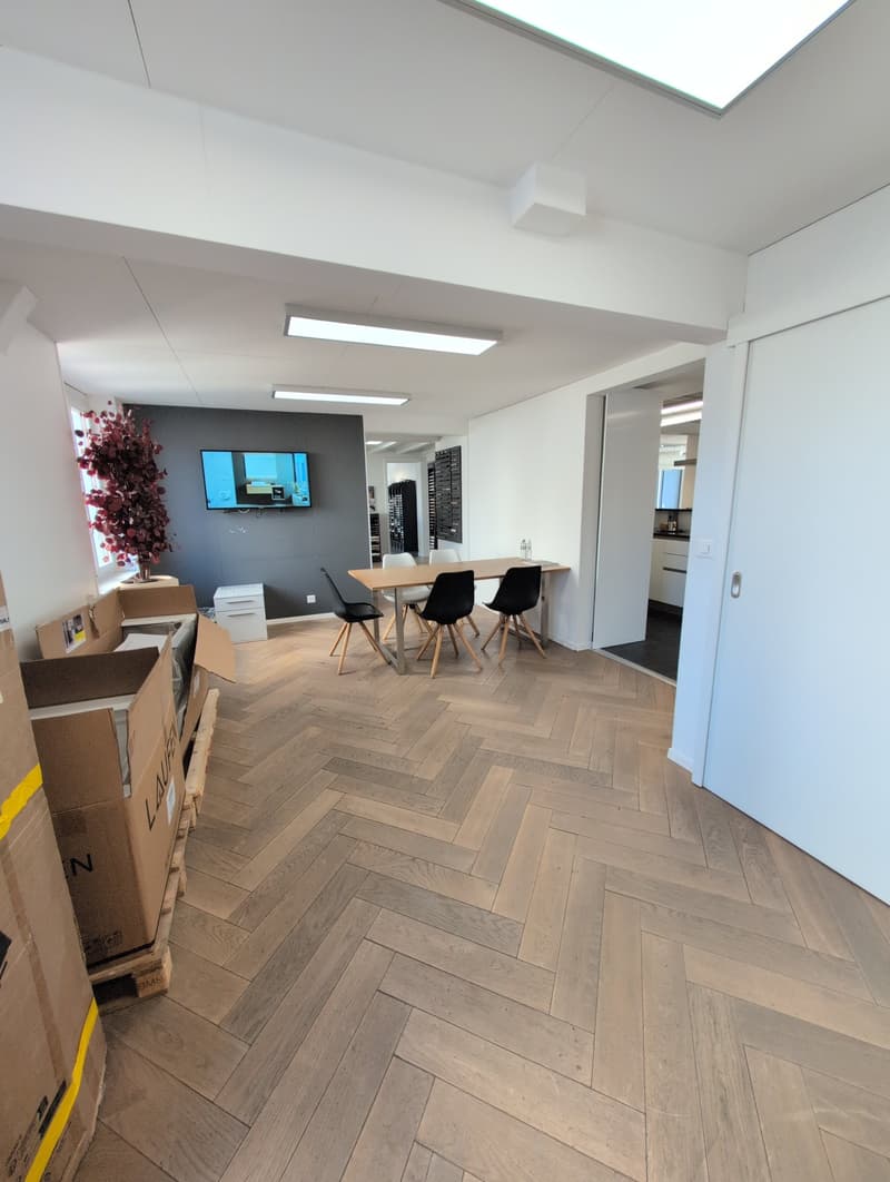 Empty room with wooden flooring, white walls, large windows, flat-screen TV mounted on wall, dining table and chairs, storage boxes, illuminated by ceiling lights.