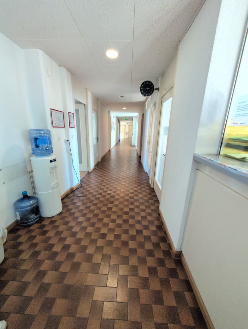 Corridor: checkered floor, white walls, ceiling, clock, water dispensers, water bottles, exit sign, open doors