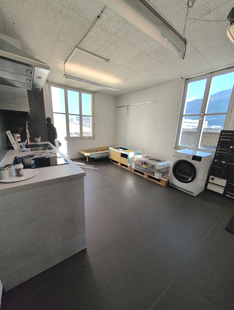 A bright kitchen with white walls and gray tiled floors, it includes a modern kitchen island with a built-in sink and a white washer placed next to a large window. Various appliances and objects are scattered around the room.