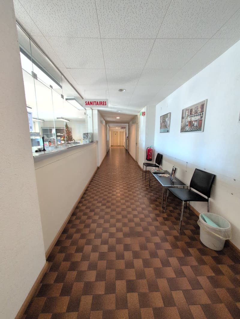 Empty hallway with checkered flooring, white walls, glass walls, two tables with chairs, fire extinguisher, trash bin, reception desk with a monitor, Christmas tree and cabinets.