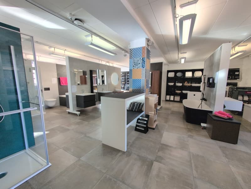 A showroom with tiled flooring and modern bathrooms exhibits, featuring a glass shower enclosure, a toilet, a white bathtub, sinks, and a central display table.