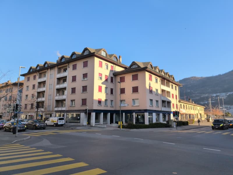 The exterior of a multistory building with glass windows, balcony, parking lot, traffic lights, and a mountain on the horizon
