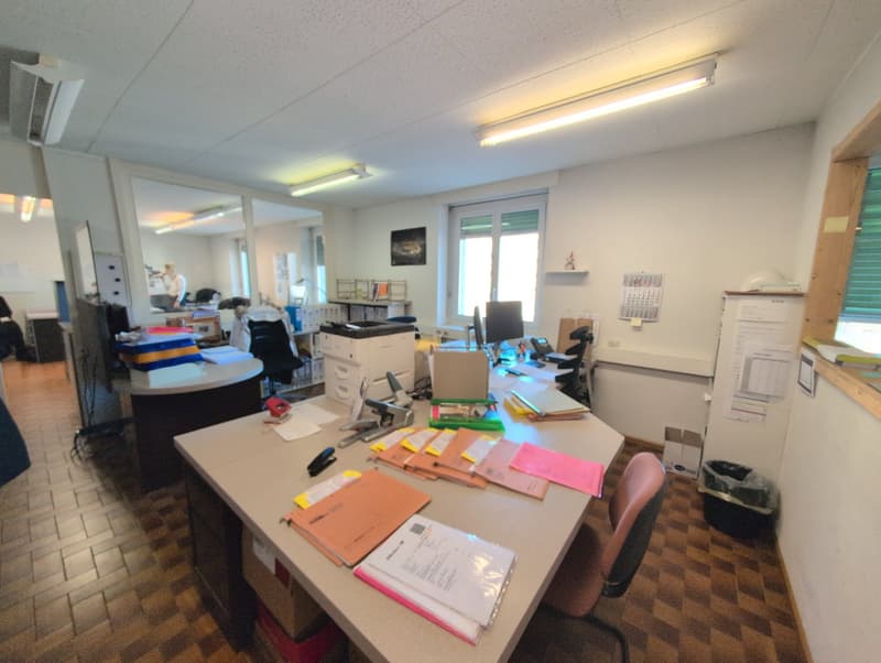 Modern office room with large windows, desk, shelves, computer monitor, office chairs, storage boxes, and a trash bin. The room has a ceiling with LED lighting fixtures, a white and wooden flooring, and a mirror mounted on the wall.